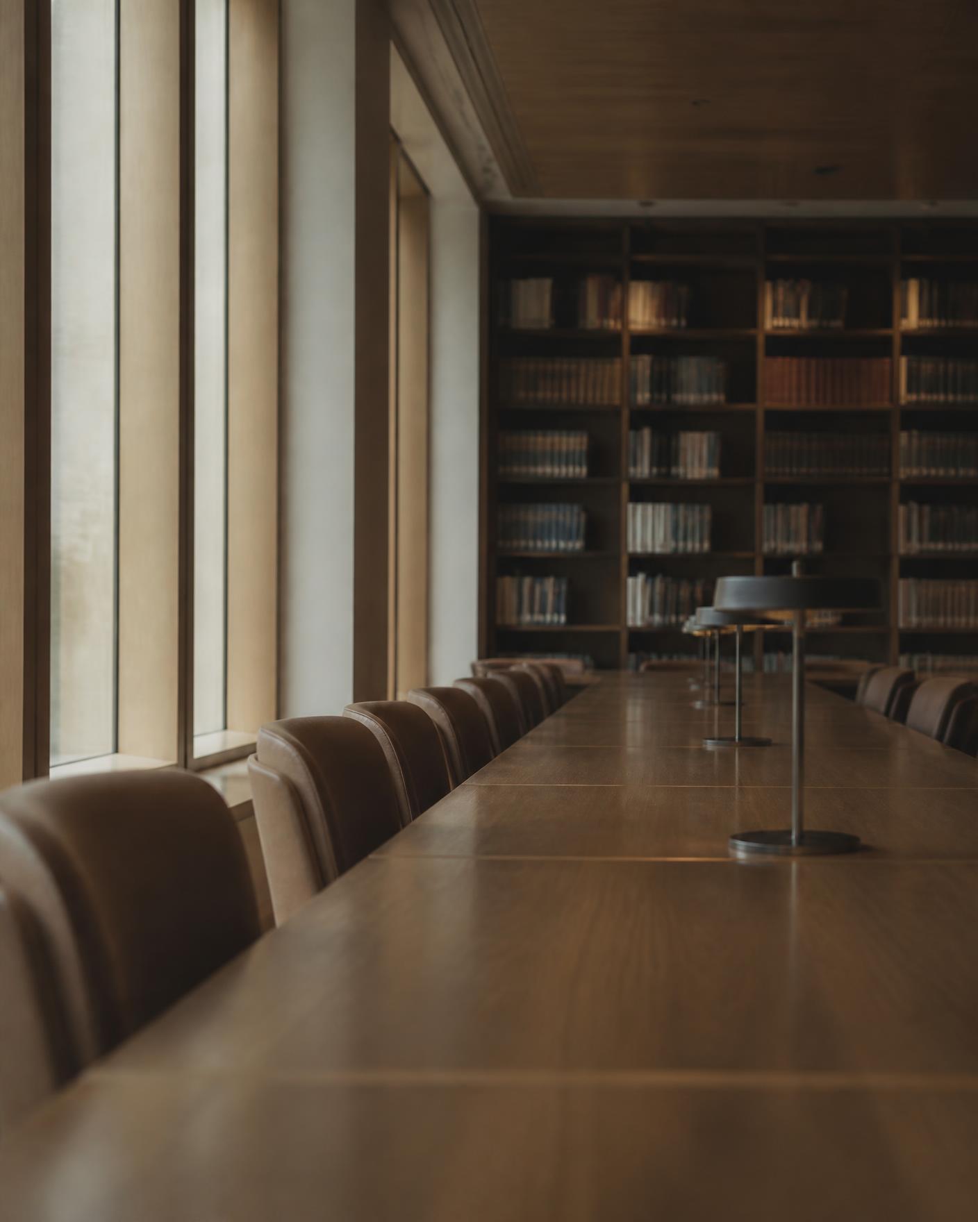 Empty reading room in a research library, long table lit by daylight from tall windows.